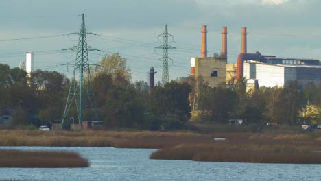Distant-view-of-abandoned-metal-melting-factory-red-brick-chimneys-and-electricity-lines-in-sunny-autumn-day,-wide-shot-from-a-distance-over-the-lake-Liepaja