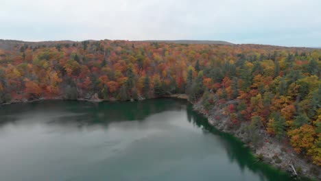 Lowering-and-panning-left-aerial-footage-over-Pink-Lake-in-Gatineau-Quebec-with-boardwalks-and-lookouts-in-view-on-the-side-of-a-hill-surrounded-by-an-autumn-forest