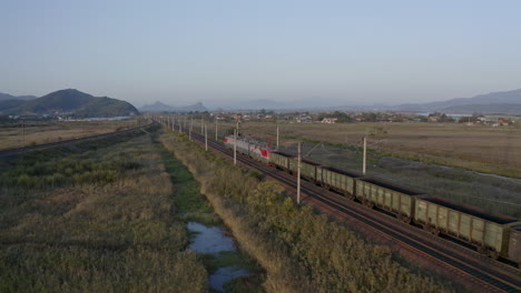 Russian-railway-train-locomotives-pulling-a-long-empty-coal-carriages-freight-through-green-fields-with-mountains-in-the-background-on-the-sunset,-Russian-Federation
