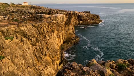 The-sun-sets-over-the-rugged-cliffs-of-Boca-do-Inferno-in-Cascais,-casting-a-warm-glow-on-the-rocks-and-ocean-below