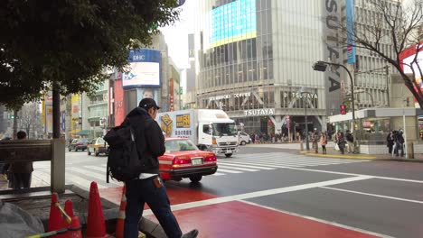 Ein-Mann-Mit-Hut-Und-Rucksack-Dampft-Vor-Der-Straßenkreuzung-In-Shibuya,-Während-Der-Verkehr-Vorbeizieht