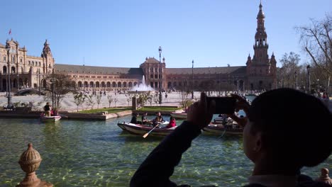 Silhouette-Asian-man-takes-picture-of-boats-in-Plaza-de-Espana-canal,-Seville