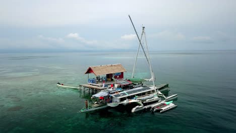 Bigkanu-trimaran-sailboat-at-an-ocean-cottage-with-man-diving-to-the-waters-from-the-ship,-Aerial-circle-around-shot