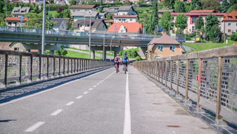 La-Gente-Monta-En-Bicicleta-En-Un-Puente-En-Muta,-Eslovenia-Con-Una-Ciudad-Ocupada-En-El-Fondo