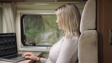 SLOWMO---Beautiful-blond-girl-sitting-in-campervan-at-table-working-on-laptop-with-trees-and-nature-in-background