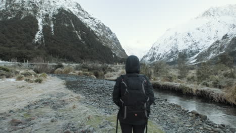 Girl-in-hiking-gear-walking-along-river-surrounded-by-snow-capped-mountains