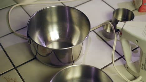 A-kitchen-counter-covered-in-metal-mixing-bowls,-measuring-cups-and-cooking-utensils-as-a-chef-prepares-to-bake-a-chocolate-cake