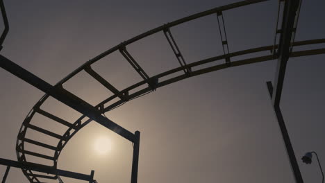Silhouette-of-a-rickety-old-roller-coaster-nearing-sunset,-camera-pans