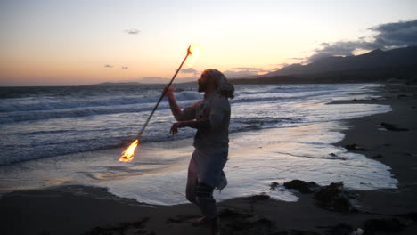A-male-flow-art-dancer-in-silhouette-performing-his-fire-spinning-on-the-beach-at-sunset-with-ocean-waves-in-slow-motion