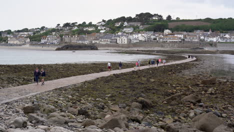 Tourists-walking-on-the-causeway-that-leads-to-Marazion-from-the-english-medioeval-castle-and-church-of-St-Michael's-Mount-in-Cornwall-on-a-cloudy-spring-day,-Zoom-out-4k-footage