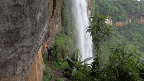Ugandan-men-hiking-on-a-muddy-trail-behind-a-large-waterfall-in-tropical-East-Africa