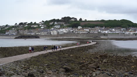 Tourists-walking-on-the-causeway-that-leads-to-Marazion-from-the-english-medioeval-castle-and-church-of-St-Michael's-Mount-in-Cornwall-on-a-cloudy-spring-day,-Zoom-in-4k-footage