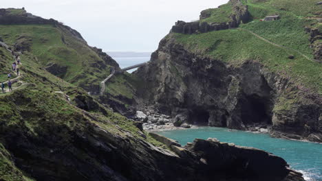 Tourists-walking-on-top-of-the-cliffs-by-the-ruins-of-Tintagel-castle-in-northern-Cornwall-with-Tintagel-beach-and-bay-below
