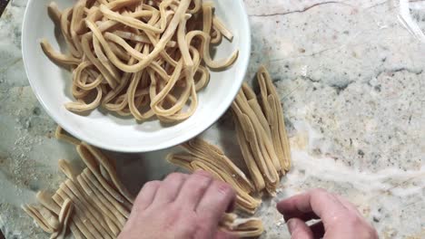 overhead-view-of-hands-opening-up-pasta-dough-noodles-and-adding-into-bowl