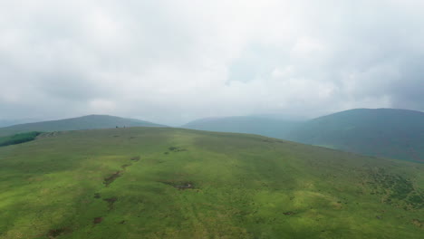 Aerial-shot-looking-over-a-grassy-hill-with-mountains-in-the-background,-overcast-day