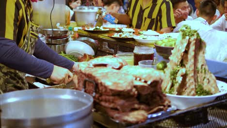 Black-and-Yellow-uniform-man-cut-meat-on-a-busy-night-food-market-stall,-Bangkok