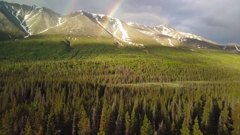 Stunning-Yukon-flight-by-double-rainbow-arc-and-picturesque-mountain-range-with-green-lush-vegetation-and-forest-trees-beneath-on-cloudy-day,-Canada,-overhead-aerial-sideways