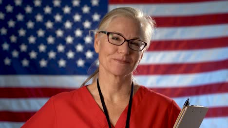 Portrait-of-a-nurse-nodding-her-head-and-smiling-against-an-out-of-focus-US-flag