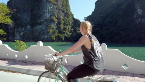 Mujer-Rubia-Con-Gafas-Montando-En-Bicicleta-En-Una-Carretera-De-La-Isla-De-Bicicletas-En-Un-Día-Soleado,-Toma-Aérea-De-Seguimiento