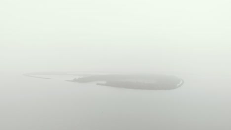 Slow-aerial-approach-of-a-mysterious-and-in-mist-banks-covered-oval-shaped-island-on-a-totally-overcast-and-grey-monotone-day-climate