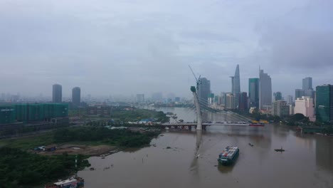 Drone-shot-of-Thu-Thiem-2-Bridge-is-a-bridge-under-construction-in-Ho-Chi-Minh-City-with-boat-carrying-shipping-containers-and-city-skylinee