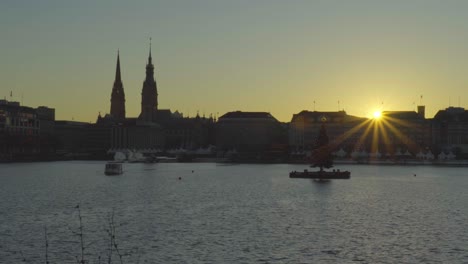View-of-the-sun-setting-on-Hamburg,-Germany,-across-Inner-Alster-Lake-with-a-tour-boat-passing-by-a-floating-Christmas-tree-in-Dec-2019