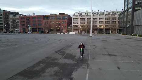 Tracking-a-young-man-on-an-electric-rental-scooter-riding-through-an-empty-parking-lot,-aerial