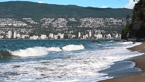 Big-waves-rooling-at-a-sandy-beach-while-white-rooling-rewinding-back-to-sea-in-Stanley-park-in-Vancouver-with-building-in-the-background-on-a-partly-cloudy-summer-day