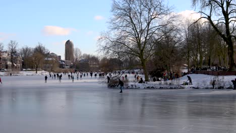 Left-panning-showing-picturesque-scenery-of-people-ice-skating-and-enjoying-on-frozen-canal-in-city-environment-with-winter-barren-trees-and-white-snow-in-cold-cozy-landscape