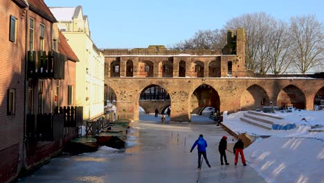 Berkelpoort-stairs-covered-in-snow-and-youngsters-making-an-ice-hockey-stick-while-an-ice-skater-passes-by-on-frozen-river-passing-underneath-the-medieval-boat-portal-city-wall