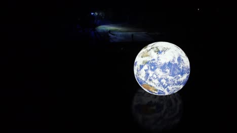 Luke-Jarram-Floating-Earth-illuminated-planet-reflecting-in-lake-water-rippling-surface-at-night-orbiting-left