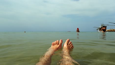 Low-angle-personal-perspective-of-male-legs-and-feet-relaxing-while-floating-on-seawater-with-people-and-trabocchi-in-background
