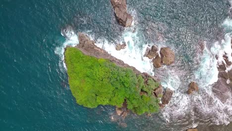 Drone-shot-of-Coral-island-overgrown-by-green-trees-that-are-buffeted-by-the-waves-crashing-in-the-tropical-ocean