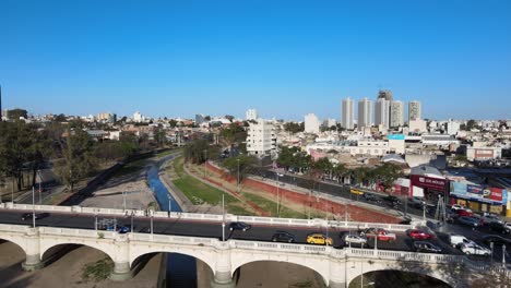 Aerial-drone-flyover-Puente-Centenario-over-Suquia-river-capturing-morning-traffics-on-arch-bridge-and-boulevard-mitre-with-downtown-cityscape-in-the-background