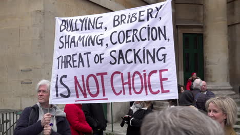Two-people-hold-a-white-banner-that-reads-in-black-and-red-writing,-“Bullying,-bribery,-shaming,-coercion,-threat-of-sacking-is-not-a-choice”-on-a-protest-opposing-mandatory-Covid-vaccinations