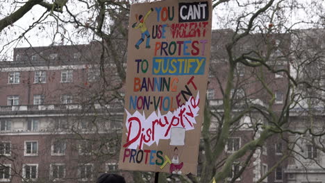 People-walk-past-a-colourful-handmade-cardboard-placard-being-held-up-that-reads,-“You-can’t-use-violent-protest-to-justify-banning-non-violent-disruptive-protest”