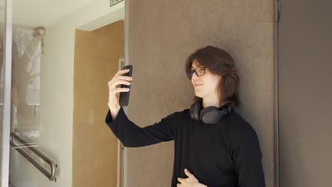 Young-man-with-glasses-on-a-video-call-using-his-cell-phone,-headphones-around-his-neck