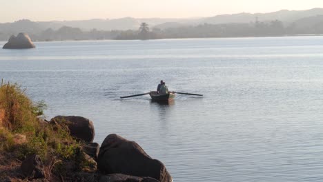 Two-men-rowing-on-a-boat-on-a-lake-during-sunrise-with-rocks-in-the-background-and-foreground-in-Aswan,-Egypt