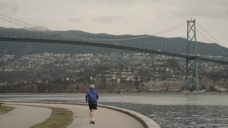 Wide-panning-shot-of-running-man-in-Stanley-park-with-Lions-Gate-bridge-in-back