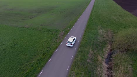 Toma-Aérea-De-Un-Dron-Con-Perspectiva-De-Pájaro-De-Un-Coche-Blanco-Conduciendo-Por-Una-Estrecha-Carretera-Rural-En-El-Sur-De-Suecia-Skåne