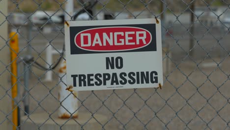 A-close-up-shot-of-a-danger-and-no-trespassing-sign-attached-to-a-chain-linked-fence-with-cars-driving-in-the-background