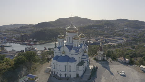 Slow-descent-aerial-shot-of-an-orthodox-church-with-blue-roof-and-golden-domes,-located-on-top-of-the-hill-with-the-reveal-of-port-and-city-building-in-the-background,-on-a-bright,-clear,-sunny-day