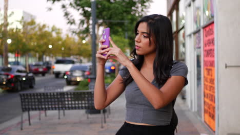 Eine-Attraktive-Junge-Hispanische-Frau,-Die-Durch-Die-Straßen-Der-Stadt-Läuft-Und-Mit-Ihrem-Smartphone-In-Zeitlupe-Fotografiert