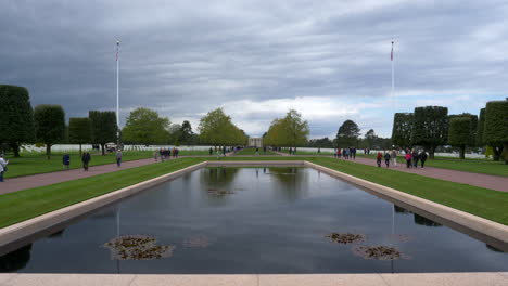 Crowd-walking-around-the-Normandy-American-Cemetery-and-Memorial-reflecting-pool,-American-flag-poles-on-either-side