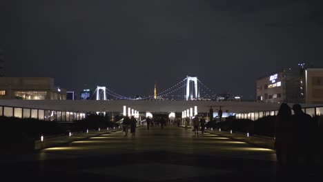 Night-scene-of-Odaiba-walkway-with-people-walking-by-and-the-Rainbow-Bridge-in-Tokyo-Bay-in-the-background,-Tokyo,-Japan