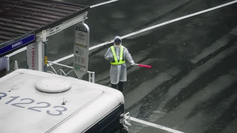 Japanese-Traffic-Guard-Wearing-Raincoat-Stopping-A-Bus-At-The-Intersection-In-Tokyo,-Japan-On-Rainy-Season---high-angle,-slow-motion