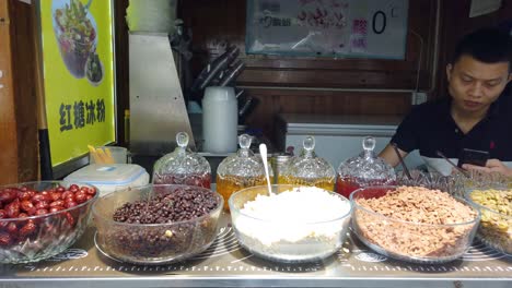 Street-stall-on-a-food-market-in-China-with-display-of-glass-bowls-filled-with-various-ingredients-like-nuts,-sweets,-figs-and-sauces