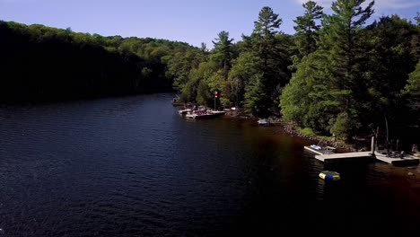 Drone-Flying-over-Cottage-Docks-on-Lake-with-Canada-and-Germany-Flags-Flying-in-Wind-and-Green-Pine-Trees-on-Shore