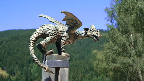 Bronze-dragon-statue-on-pole,-mountain-landscape,-forest-and-alpine-pasture