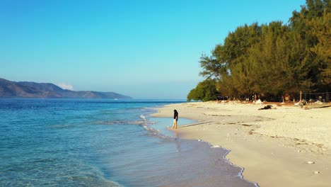 Lonely-girl-standing-on-wet-sand-of-exotic-beach-in-front-of-white-waves-coming-from-blue-sea-in-coastline-of-tropical-island-of-Indonesia